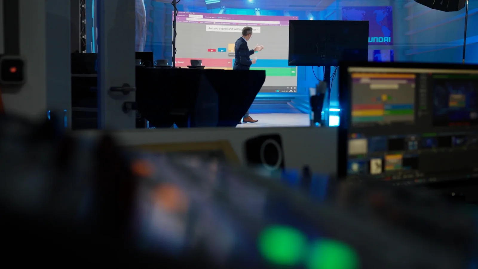 Blick vom Regieplatz über Mischpult und Kontrollmonitore auf die Stage, auf der LED-Wand läuft eine Live-Umfrage mit Antwortbalken